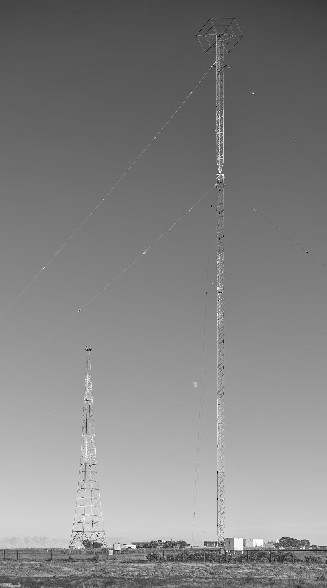 Two radio antenna masts in a field. The taller mast, on the left, is a guyed tower with a hexagonal structure at the top. The shorter mast on the right, toward the background, is freestanding.