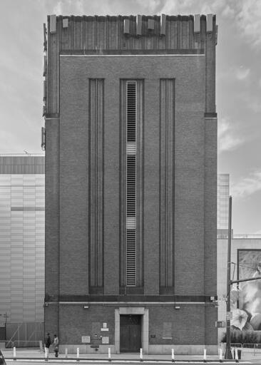 A simple art deco style building, lacking windows, but with a narrow louvered vent running vertically along its length, on a city street.
