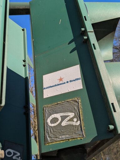 An einer Ampel kleben seitlich zwei Sticker. Auf dem ersten steht "Antifaschismus & Graffiti", auf dem zweiten "OZ we miss you"