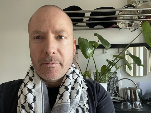 Me (white passing guy with short hair and dirty beard) standing in our hallway wearing my keffiyeh (traditional Palestinian white scarf with black motif and grey shading in parts) over a black t-shirt, in front of a plant, small mirror, some caps on a shelf, a square black abd white illustration of leaves, a metal watering can and a pair of glasses.