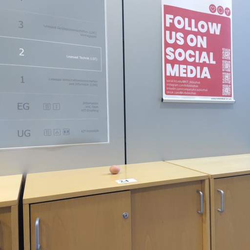 Auf einem Rollschrank in der KIT-Bibliothek liegt ein einzelnes Ei. Zu sehen ist neben dem "Stockwerke"-plan das "Follow us on Social Media"-Plakat der Bibliothek, wo neben deren Insta, linkedin und tiktok auch der mastodonkanal aufgeführt ist
