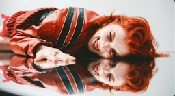 A person with bright red hair is lying on a reflective surface, displaying an intense facial expression. They are wearing a red jacket with black stripes, and their hand rests on the surface, capturing their reflection.
