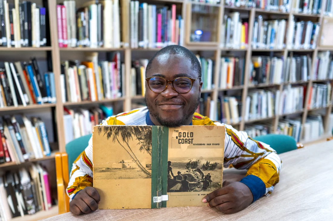 Image of Paul Ninson, a Black man, who is sitting in front of shelves of books and holding up a book entitled "The Gold Coast Yesterday and Today," which was published in the early 1940s.