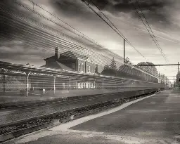An early 20th century passenger train station, with an awning over the platform, across tracks. A blurred train streaks by in foreground.