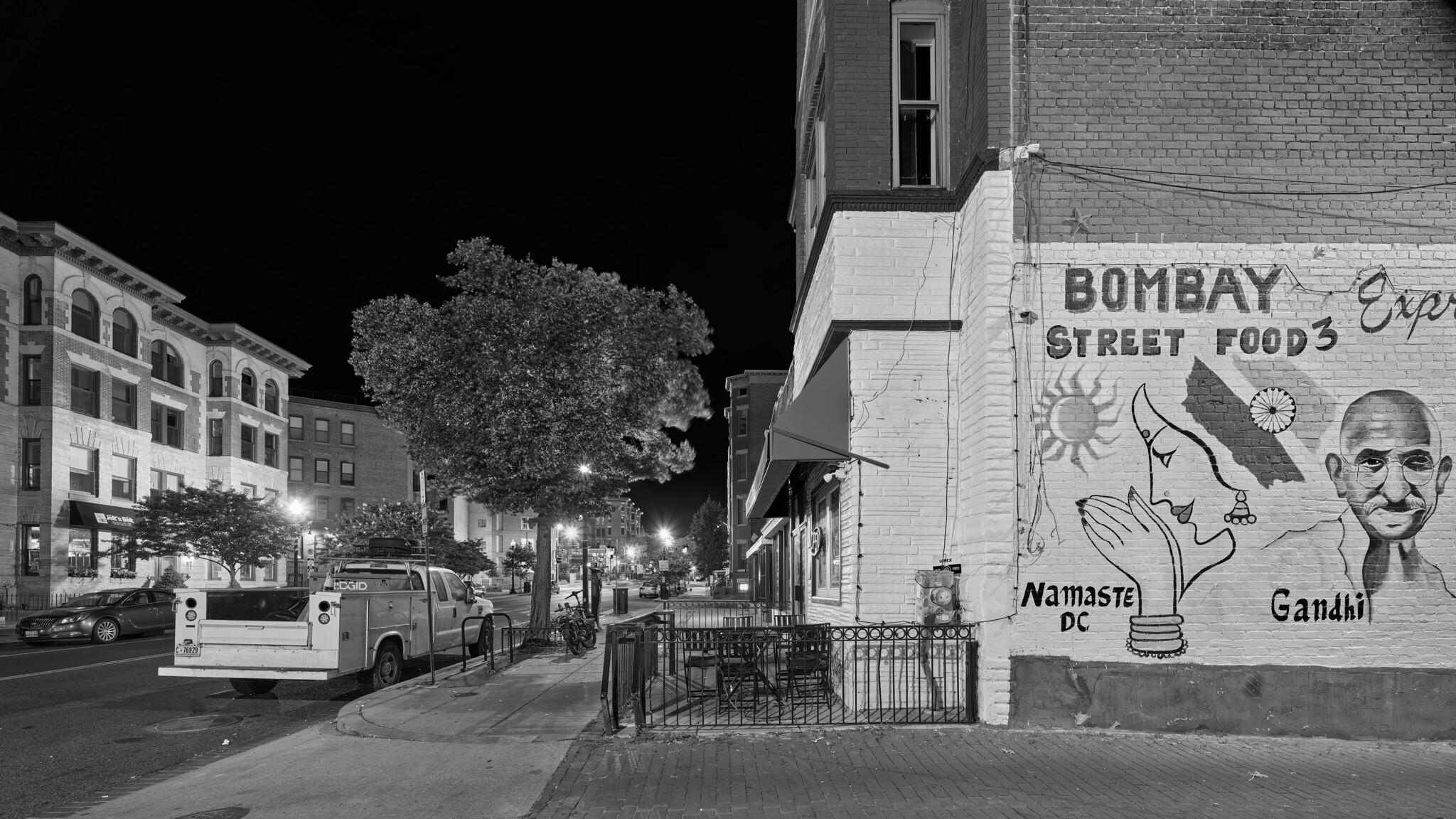 An urban street at night, devoid of people. Across the street at left, small apartment buildings. At right, a small restaurant with a mural reading "Bombay Street Food 3".