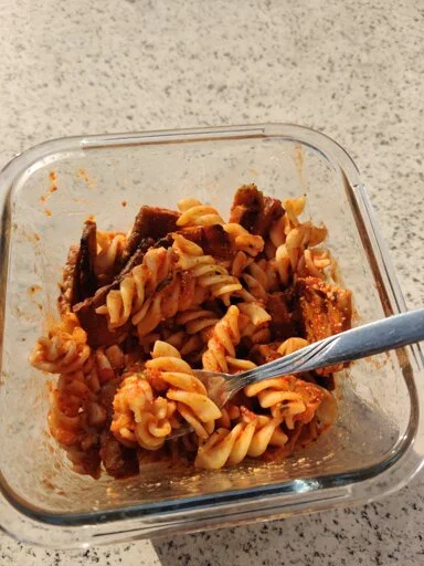 Photo of noodles (Fussili) in with sauce and fried chickpea-blocks in a glass container.