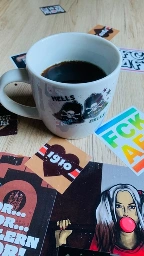 A white coffee mug filled with black coffee sits on a wooden surface, surrounded by various colorful stickers. The mug features skull and bell graphics. Stickers include playful phrases and designs, with notable images and bold colors.