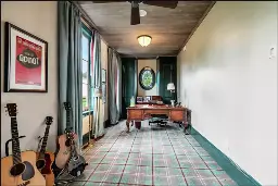 Long office with plaid carpet. On the left wall stands 3 acoustic guitars and a mandolin. Above the instruments is a poster from the Broadway production of "Waiting for Godot" starring Nathan Lane & John Goodman. Green curtains decorate two wall-sized pane windows. At the end of the office is an oval paned window and a handsome carved oak desk. Above it all is a ceiling fan and a domed light fixture above the desk.