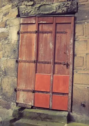An old brown wooden door with visible metal strap hinges. The bottom of the door is patched with four wooden squares painted red-orange. Marburg, Germany. Photo by Peachfront. 2008.