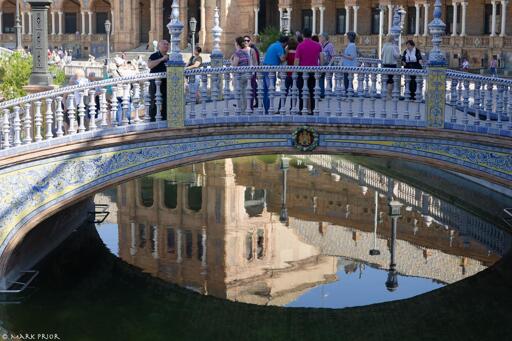 Reflection of the surrounding building in the canal under the bridge of the Kingdom of Castile at the Plaza de España in Sevilla.
A pedestrian bridge gently arches across a canal. The bridge is faced with blue and yellow tiles with a floral motif. There are a series of sculpted blue tiled pillars making up the balustrade of the bridge. In the centre of the span is the badge of the ancient Kingdom of Castile. People are standing on the bridge admiring the tile work and the surrounding building.
Under the bridge is a reflection in the canal of one of the towers of the encircling building.