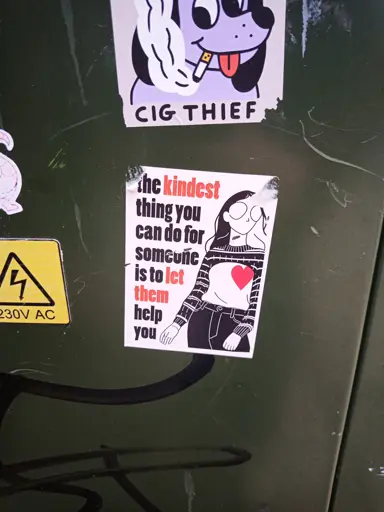 A sticker on a green utility box that has an illustration of a person with a long sleeve jumper rolled up above their chest and a red heart overlayed over one side of their chest. it has text next to it reading "the kindest thing you can do for someone is to let them help you"