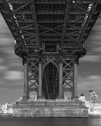 Underside of an early 20th century bridge, exposing girders and steelwork, over a river, at night.