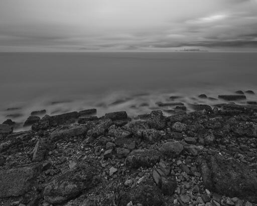 A rocky shoreline with gentle waves lapping at it. In the distance at right is a small island, somewhat enveloped by fog, with a prominent water tower and industrial-type buildings.