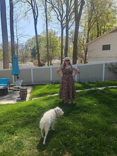 I am standing in my back yard, on the grass. Behind me is a patio with furniture. I am wearing a strappy dress with sweetheart neckline and ruffled hem on the A line skirt, over a pale blue tee. The print on the dress is woodland animals (fox, deer, owl, squirrel, moths) and trees on a navy background 